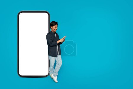 Téléchargez les photos : Heureux homme asiatique millénaire debout et utilisant un téléphone avec un énorme écran vierge smartphone maquette à côté de lui sur un fond turquoise lumineux, illustrant la connectivité et l'espace pour le design - en image libre de droit
