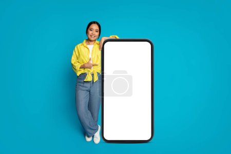 Téléchargez les photos : Femme japonaise millénaire joyeuse en chemisier jaune et jean appuyé sur un grand smartphone vertical avec un écran vierge, parfait pour le contenu publicitaire, sur un fond bleu vif - en image libre de droit