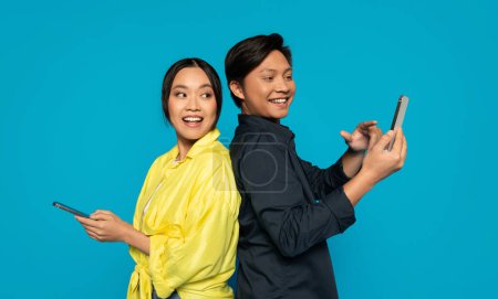 Téléchargez les photos : Deux individus asiatiques millénaires dos à dos en tenue décontractée, homme souriant en utilisant son téléphone, femme à l'expression surprise tenant également un téléphone, sur un fond turquoise vif - en image libre de droit