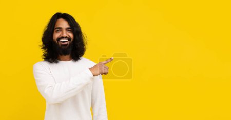 Photo for Cheerful bearded long-haired young indian man in white long sleeve pointing at empty copy space mockup for advertisement, isolated on yellow studio background, web-banner for online offer - Royalty Free Image