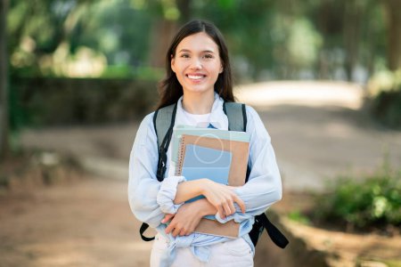 Photo for Cheerful european student college student lady with a radiant smile carrying textbooks and a backpack while walking through a lush green park on a sunny day, outdoor. Study, education - Royalty Free Image