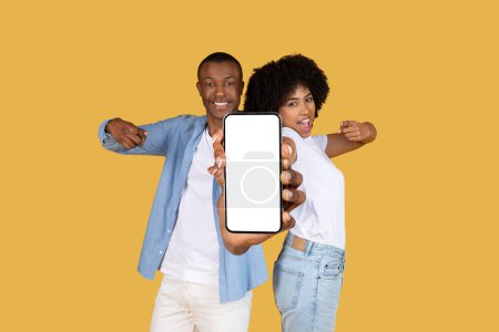 Téléchargez les photos : Heureux couple afro-américain millénaire excité présentant un smartphone avec un écran vide vers la caméra, idéal pour la promotion d'applications ou de sites Web, sur un fond jaune joyeux - en image libre de droit