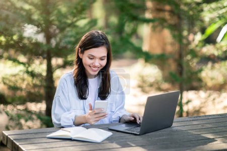 Photo for Contented european young woman student multitasking with a smartphone and laptop, sitting at a wooden table outdoors, surrounded by lush greenery in a peaceful park setting - Royalty Free Image