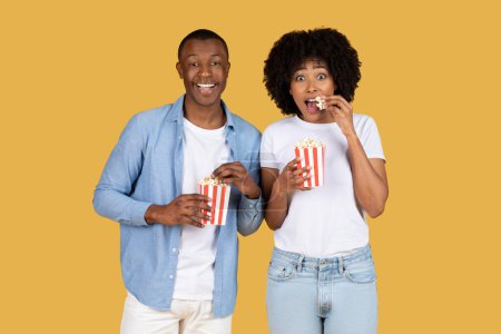 Téléchargez les photos : Joyeux couple afro-américain millénaire profitant du pop-corn, partageant un moment lumineux avec des sourires lumineux, symbolisant les loisirs, le divertissement et les joies simples de la vie - en image libre de droit