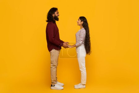 Foto de Un hombre y una mujer de pie tomados de la mano, uno frente al otro sobre un fondo amarillo vibrante, retratando una sensación de conexión y unión - Imagen libre de derechos
