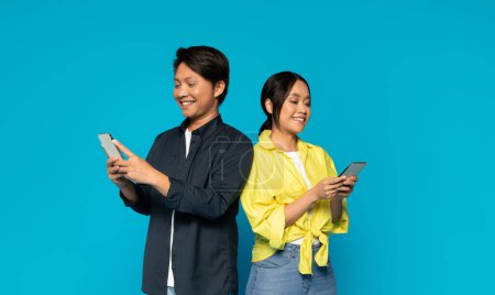 Photo for Content millennial japanese man and woman stand side by side, each happily using their smartphones, clad in a dark jacket and a yellow top respectively, against a vivid cyan background - Royalty Free Image