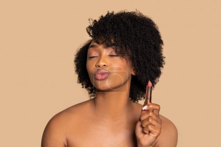 Téléchargez les photos : Femme noire avec les cheveux bouclés tient avec confiance rouge à lèvres, rondeur ses lèvres avec une expression ludique, incarnant la beauté et le plaisir sur fond beige - en image libre de droit