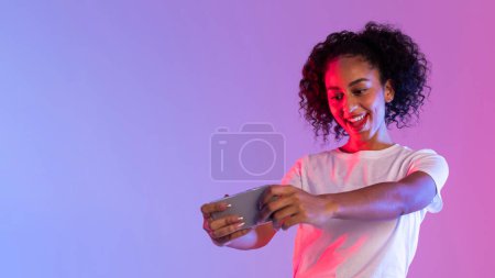 Aufgeregte Frau, die sich beim Spielen vor neonrosa und lila Hintergrund auf ihr Smartphone konzentriert