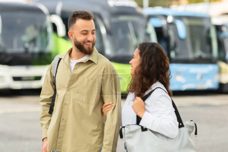 Lächelndes junges Paar, das mit Taschen am Busbahnhof spaziert und plaudert, glückliche Millennials, die ihre gemeinsame Zeit genießen, liebende Ehepartner, bereit für Reisen und neue Abenteuer, freier Raum
