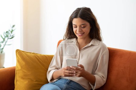 Photo for Cheerful young woman in casual shirt browsing social media on her smartphone while sitting comfortably on couch at home, happy female relaxing with mobile phone on sofa in living room, copy space - Royalty Free Image