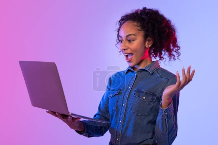 Enthusiastic woman with curly hair and arms spread wide using a laptop, pink and blue backdrop