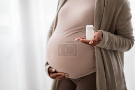 Photo for Unrecognizable pregnant black woman embracing her big tummy, holding jar with pills drugs medication, standing next to window at home, copy space, cropped. Supplements for future moms - Royalty Free Image