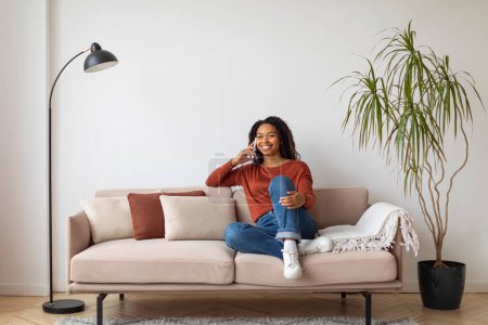 Photo for Relaxed young black woman enjoying conversation on her mobile phone while comfortably lounging on beige couch at home, smiling african american lady talking on cellphone in living room, copy space - Royalty Free Image