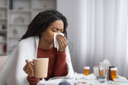 Téléchargez les photos : Jeune femme noire souffrant de rhume, se mouchant et tenant une tasse de thé, femme afro-américaine malade assise à la maison entourée de médicaments, montrant des symptômes de grippe, espace de copie - en image libre de droit