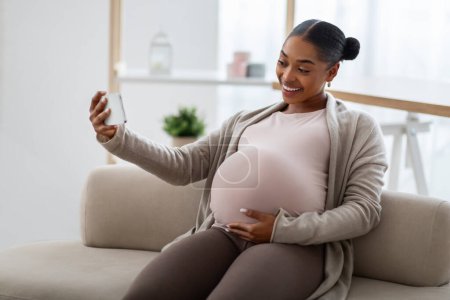 Fröhliche afrikanisch-amerikanische junge schwangere Frau, die zu Hause auf der Couch sitzt, ihr Smartphone benutzt, sich online mit Ehemann oder Freund trifft und ihren dicken Bauch zeigt