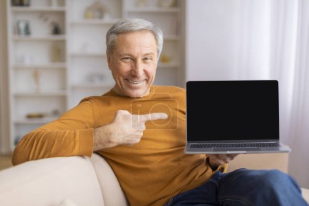 Téléchargez les photos : Homme âgé assis sur un canapé, pointant vers l'écran vierge d'un ordinateur portable, parfait pour la publicité ou des messages éducatifs - en image libre de droit