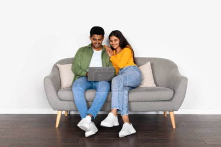 Fröhliches indisches Paar mit Laptop sitzt gemütlich auf modernem Sofa und teilt lustige Momente in stilvollem, gut beleuchtetem Wohnraum mit weißer Wand, volle Länge