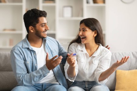 Ekstatische junge indische Ehepartner mit positivem Schwangerschaftstest, die auf der gemütlichen Wohnzimmercouch fröhlich Zukunftspläne mit animierten Gesichtsausdrücken diskutieren