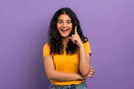Téléchargez les photos : Femme joyeuse en t-shirt jaune avec eureka moment, pointant vers le haut, debout sur un fond violet - en image libre de droit
