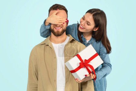 Photo for Young woman covers her boyfriends eyes surprising him with a birthday present against blue backdrop, posing with wrapped gift box, celebrating Valentines day or romantic relationship anniversary - Royalty Free Image