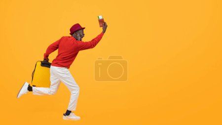 Téléchargez les photos : Homme noir sénior excité dans un pull rouge et chapeau fonctionnant avec un passeport et une valise jaune, montrant le mouvement dynamique et la joie de voyager sur un fond jaune vif, studio - en image libre de droit