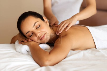 Photo for Spa and relaxation. Smiling young lady enjoys a professional back massage indoors at beauty center, for comfort and self care, lying wrapped in towel while masseuse massaging her back muscles - Royalty Free Image