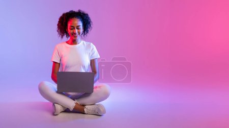 Téléchargez les photos : Une femme souriante aux cheveux bouclés utilise un ordinateur portable assis avec les jambes croisées sur un fond dégradé - en image libre de droit