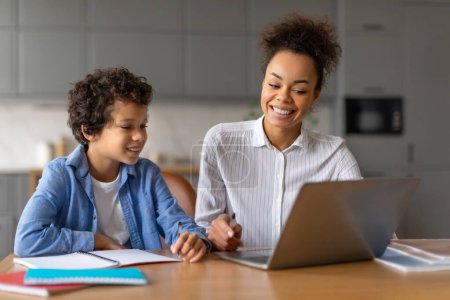 Téléchargez les photos : Une mère et son jeune fils sourient alors qu'ils sont assis à une table travaillant ensemble sur un ordinateur portable, ce qui implique une scolarité à domicile ou une session de devoirs - en image libre de droit