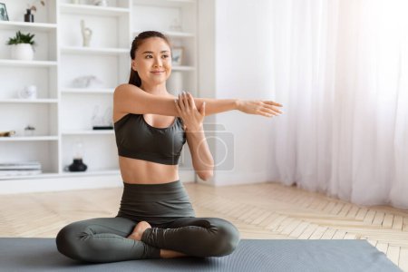 Foto de Mujer asiática sonriente en ropa de yoga que realiza ejercicio de estiramiento del brazo sentado, hermosa mujer coreana deportiva que se ve relajada mientras entrena en el interior de la sala de estar brillante, practicando yoga en casa - Imagen libre de derechos