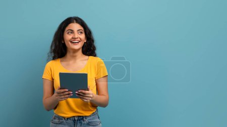 Eine fröhliche junge Frau im gelben Hemd hält ein digitales Tablet in der Hand und blickt mit freudigem Gesichtsausdruck auf blauem Hintergrund nach oben.