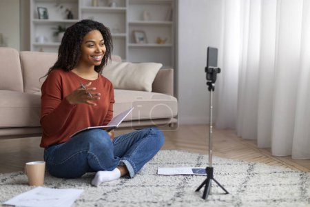 Photo for Online Coaching. Black Female Tutor Filming Video Lecture Via Smartphone At Home, Smiling African American Woman Holding Notepad, Talking And Gesturing On Mobile Camera, Enjoying E-Teaching - Royalty Free Image