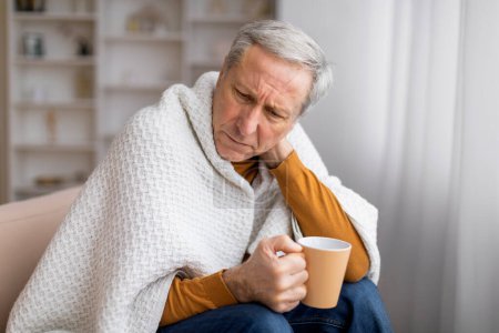 Foto de Enfermo mayor envuelto en una manta mientras sostiene una taza, sentado en el sofá, ilustrando problemas de salud o recuperación - Imagen libre de derechos