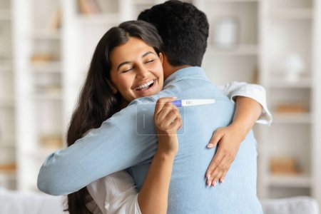 Photo for Overjoyed indian woman embracing man from behind, holding positive pregnancy test, both sharing moment of happiness and anticipation in bright room - Royalty Free Image