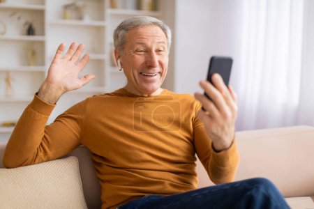 Téléchargez les photos : Heureux homme âgé utilisant un smartphone pour le chat vidéo, agitant avec enthousiasme à l'écran avec une expression joyeuse - en image libre de droit