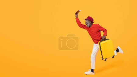 Photo for Energetic senior black man in a red sweater and hat sprinting with a yellow suitcase, holding up a boarding pass, showing the thrill of travel on an orange background, studio - Royalty Free Image