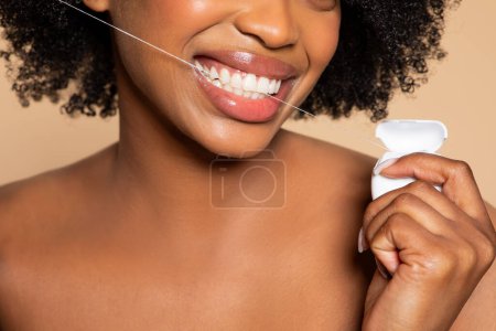 Photo for Close-up of cheerful black woman flossing her pearly white teeth with dental floss, showcasing bright, healthy smile against beige backdrop - Royalty Free Image