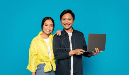 Téléchargez les photos : Couple asiatique millénaire souriant étroitement debout ensemble, avec l'homme tenant un ordinateur portable et la femme appuyée sur son épaule, sur un fond turquoise propre, suggérant la coopération - en image libre de droit