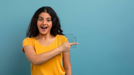 Photo for A woman in a yellow shirt points excitedly to the side, eyes wide open, with a blue background - Royalty Free Image