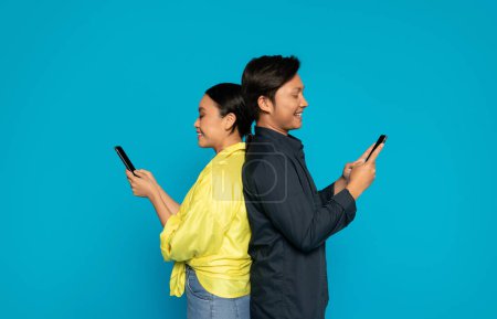 Foto de Dos millennials individuos asiáticos alegres, espalda con espalda, están sonriendo y absortos en sus teléfonos inteligentes contra un vibrante fondo turquesa, exudando un ambiente casual y conectado - Imagen libre de derechos