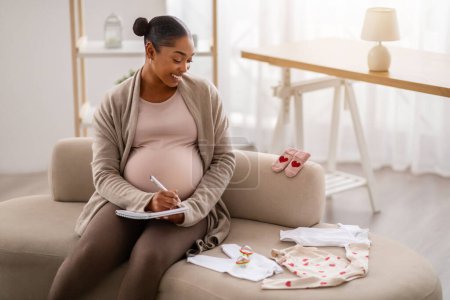 Photo for Happy young black pregnant woman with big tummy sitting on couch at home, looking at baby clothes, making list, getting ready for motherhood or labor, copy space - Royalty Free Image
