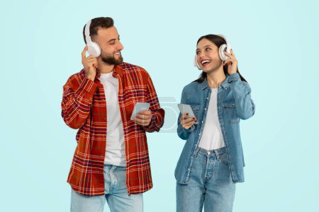 Photo for Musical Connection. Happy Young Couple In Casual Holding Smartphones Wearing Wireless Headphones, Listening To Music Online Together, Relaxing Over Blue Background, Studio Shot - Royalty Free Image