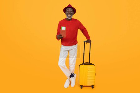 Photo for Smiling senior black man in a stylish red hat and sweater leaning on a yellow suitcase, holding a passport and boarding pass, representing joyful travel readiness on an orange background - Royalty Free Image