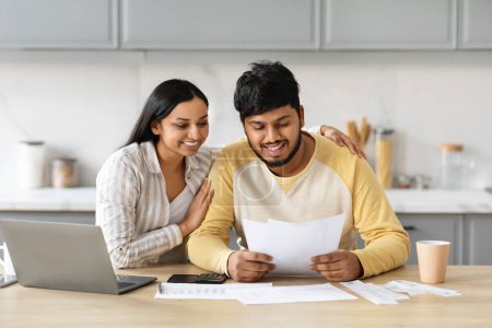 Téléchargez les photos : Conjoints indiens joyeux payant des factures sur Internet de la maison, assis ensemble à la table de cuisine, tenant des lettres et des papiers, utilisant l'ordinateur et souriant, appréciant l'e-banking facile, l'espace de copie - en image libre de droit
