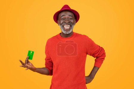 Photo for A cheerful senior black man with a bright white smile, wearing a red hat and sweater, holds a green credit card, standing against an orange background, studio. Finance, money - Royalty Free Image