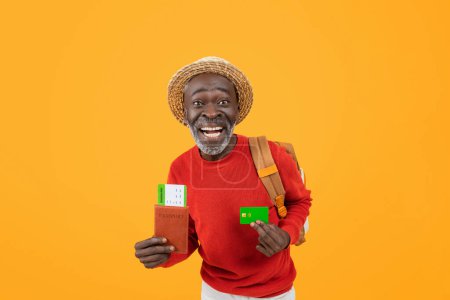 Foto de Hombre negro senior listo para viajar con una sonrisa radiante, con pasaporte y tarjeta de crédito, vestido con suéter rojo y sombrero de paja, listo para la aventura sobre un fondo naranja - Imagen libre de derechos