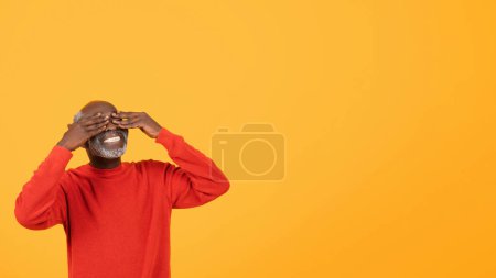 Foto de Riendo anciano negro cubriendo sus ojos con las manos, vistiendo un suéter rojo brillante, de pie felizmente sobre un fondo amarillo vivo con espacio de copia, estudio, panorama - Imagen libre de derechos