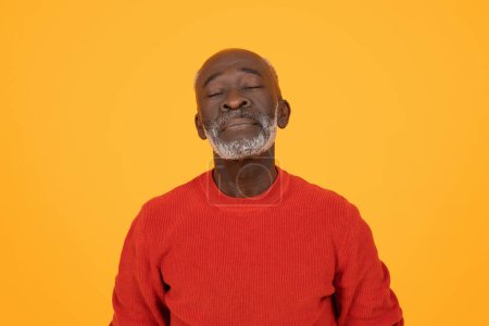 Téléchargez les photos : Contenu homme noir senior avec les yeux fermés et un sourire paisible, portant un pull rouge, respirant calme et tranquillité sur un fond orange chaud, studio. Repos, relax, pause - en image libre de droit