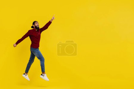 Foto de Emocionado guapo barbudo joven indio hipster saltando en el aire aislado en el fondo del estudio amarillo, apuntando al espacio de copia, mostrando una gran oferta en línea o trato increíble, longitud completa - Imagen libre de derechos