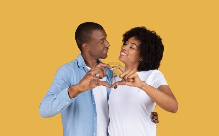 Photo for Loving millennial African American couple creating a heart shape with their hands around a house key, symbolizing love and commitment to a new home, against a vibrant yellow background - Royalty Free Image