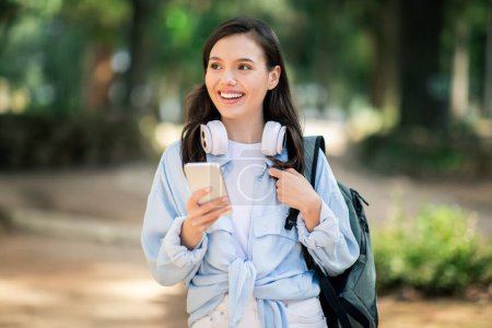 Joven estudiante europea sonriente con auriculares alrededor de su cuello usando un teléfono inteligente en un parque empapado de sol, simbolizando la conectividad y el aprendizaje moderno, al aire libre. Estudio, educación